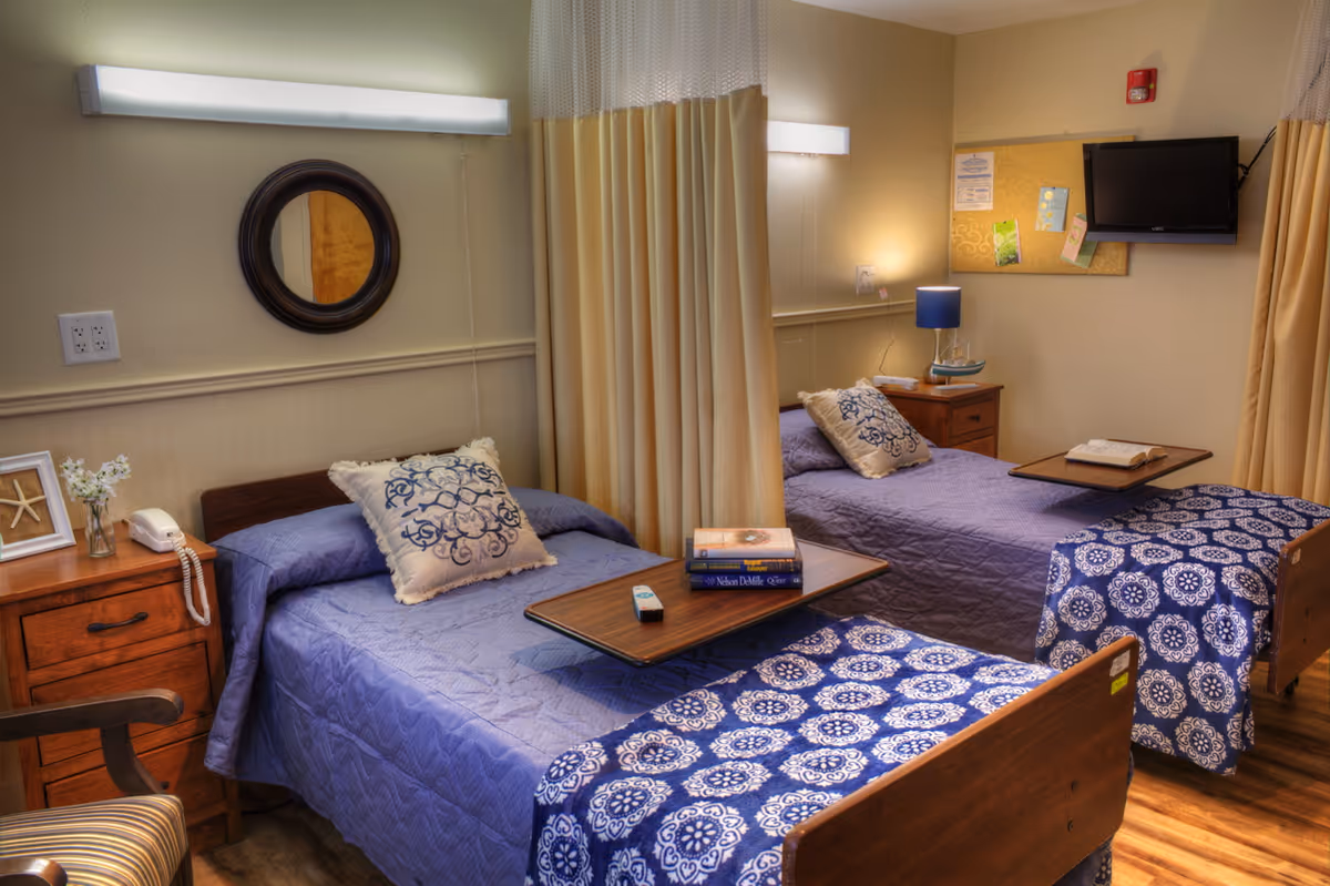 A shared bedroom in a senior living facility with two single beds separated by a privacy curtain. Each bed has a blue quilt and a decorative pillow. There are wooden nightstands beside each bed, one with a telephone and a small vase with flowers, the other with a blue lamp and some items. A small wall-mounted TV and a bulletin board with notes are visible on the wall. The room has warm lighting and wooden flooring.
