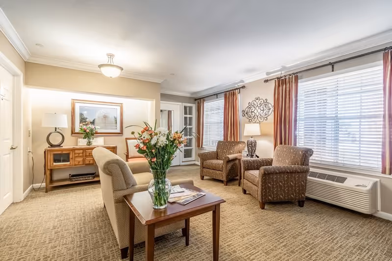 A bright and cozy living room with beige walls and carpet. The room features two patterned armchairs with a side table and lamp between them, a beige sofa, and a wooden coffee table with a vase of flowers and magazines. Large windows with white blinds and red curtains allow natural light to fill the space. A wooden console table with a lamp and framed artwork is against one wall.