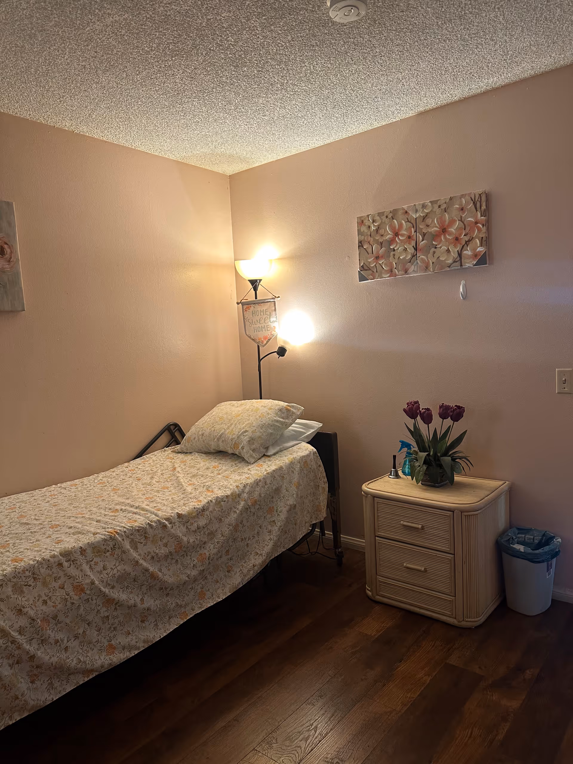 A small bedroom with a single bed covered in a floral bedspread and a pillow. Next to the bed is a light-colored nightstand with two drawers, a vase with purple flowers, a small bell, and a spray bottle on top. A floor lamp with two lights stands in the corner, one light shining upward and the other downward. The walls are painted light pink, and there are two floral paintings hanging on the walls. The floor is wooden, and there is a small trash can beside the nightstand.