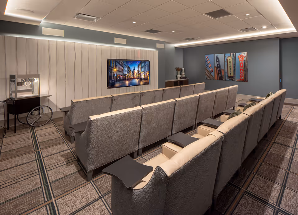 A cozy media room with two rows of cushioned theater-style seats facing a wall-mounted flat-screen TV displaying a city street scene. The room has patterned carpet, soft lighting from the ceiling, a popcorn machine on a stand to the left, and colorful artwork on the right wall.