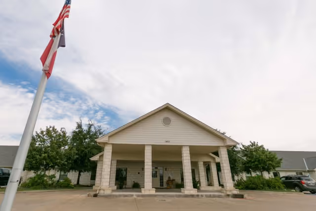 Front exterior view of Granite Mesa Health Center showing a single-story building with a covered entrance supported by stone columns, an American flag and Texas state flag on a flagpole to the left, and some trees and parked cars around the building under a partly cloudy sky.