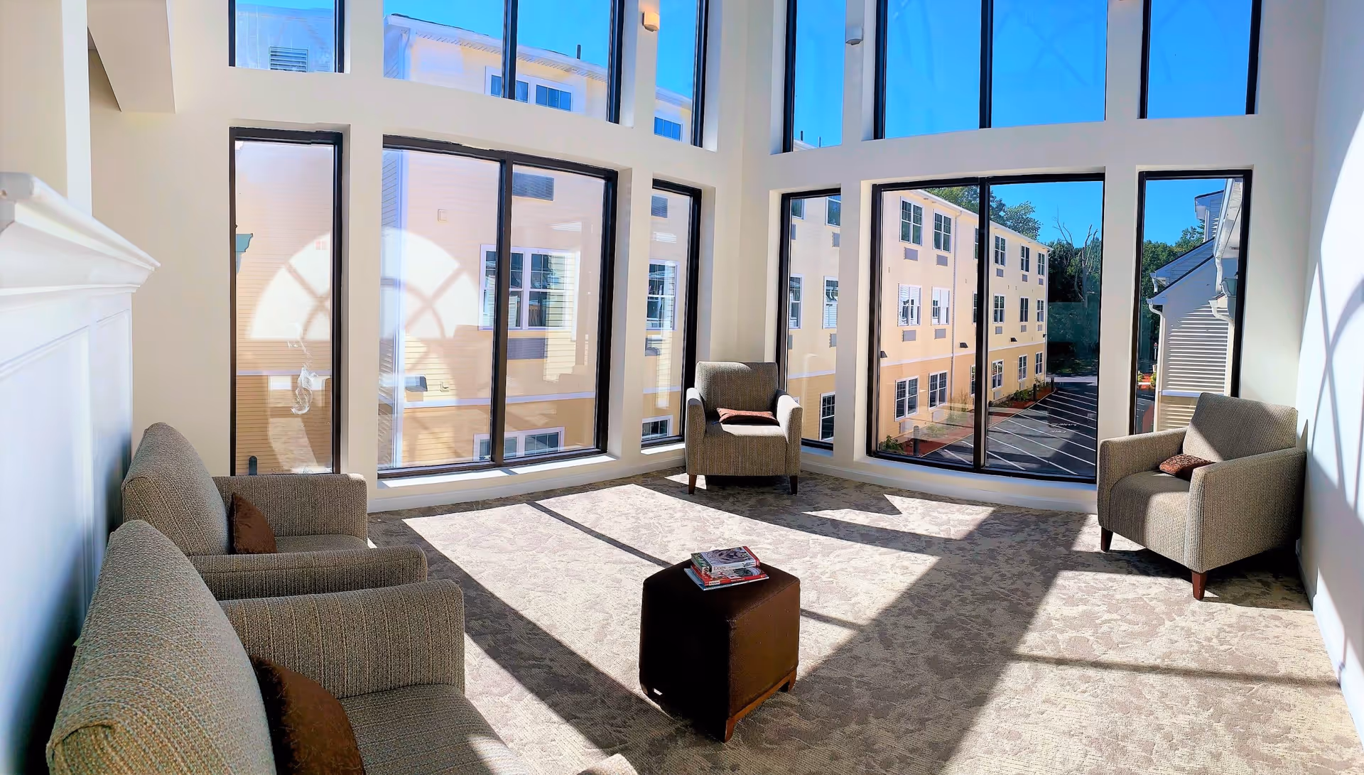 A bright and spacious sitting area with large floor-to-ceiling windows letting in natural light. The room features two armchairs and a loveseat with brown cushions, arranged around a small brown ottoman with magazines on top. Outside the windows, a parking lot and neighboring buildings are visible.