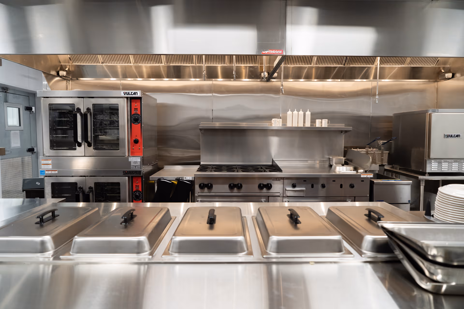 A commercial kitchen with stainless steel appliances including ovens, stovetops, and covered food trays on a counter. There are condiment bottles and cooking utensils on a shelf above the stove.