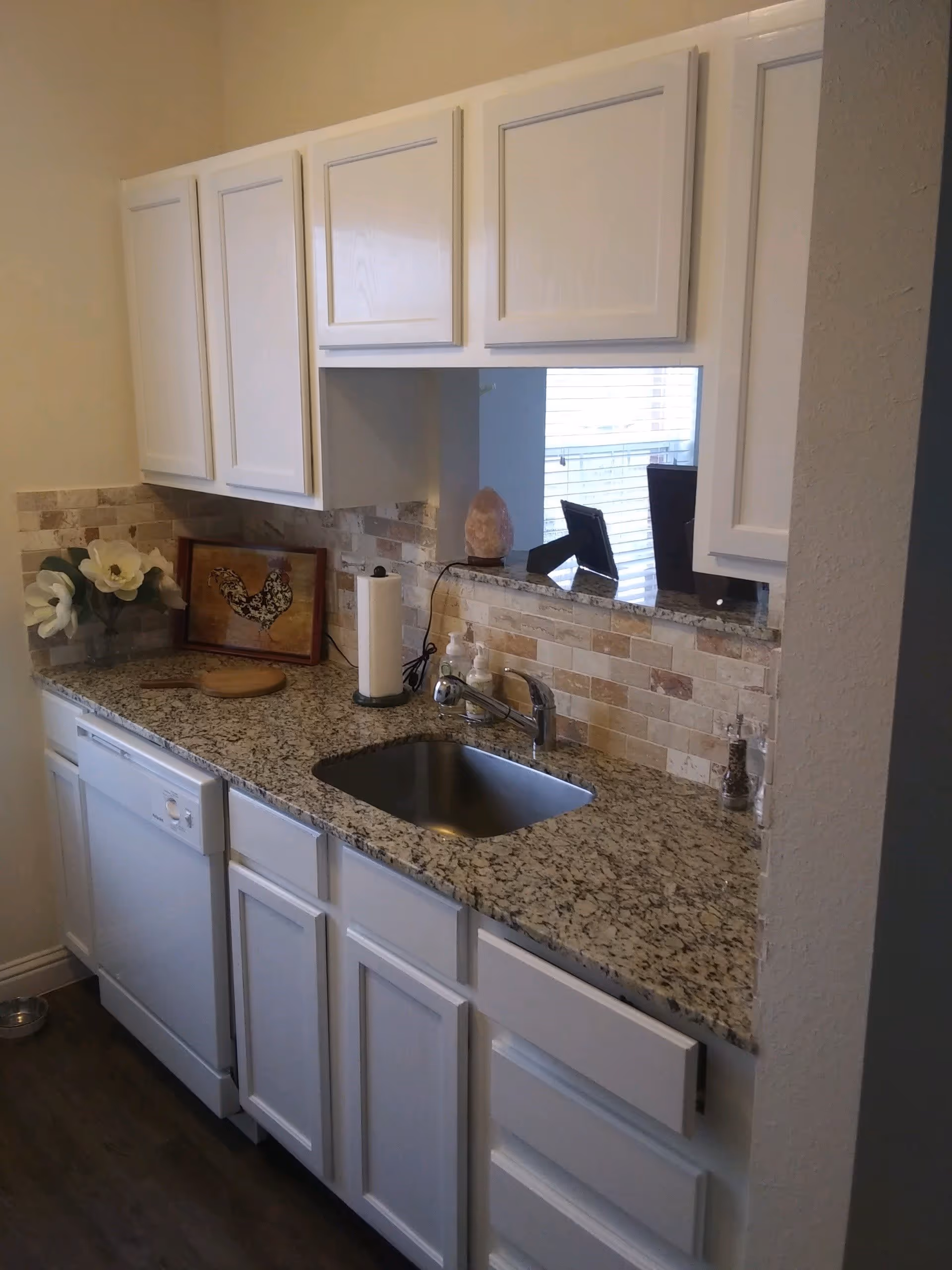 A kitchen area with white cabinets, a granite countertop, a stainless steel sink with a faucet, a dishwasher, and a tiled backsplash. On the countertop, there are decorative items including a framed picture of a rooster, a paper towel holder, a soap dispenser, and a salt or pepper grinder. A window with blinds is visible in the background.