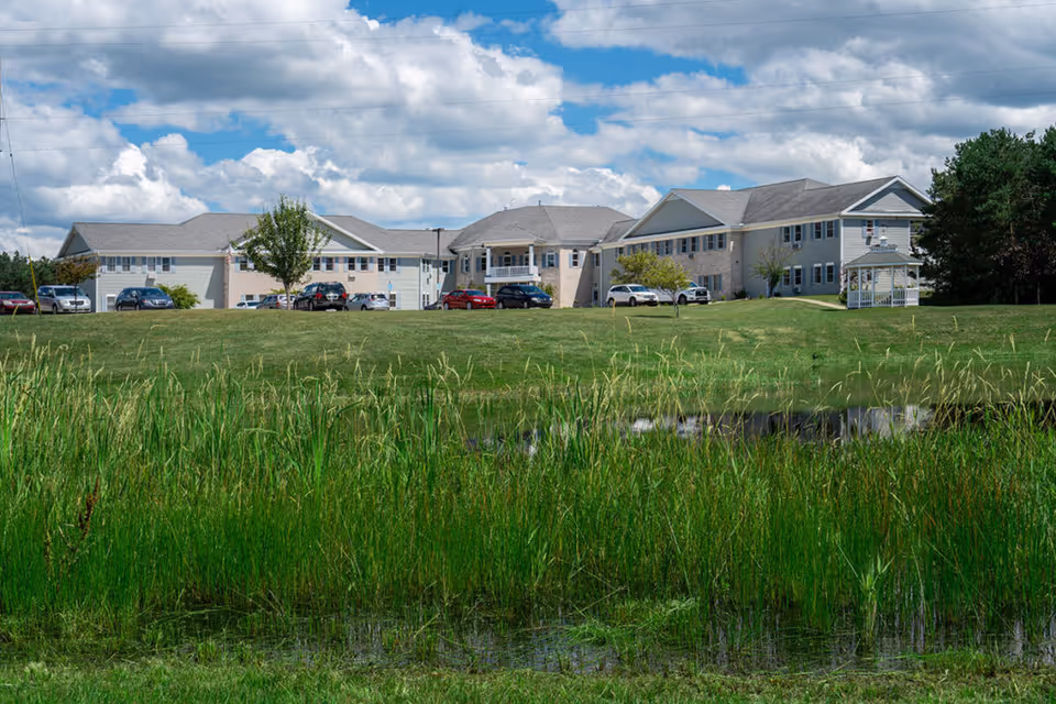 Two-story senior living building with parked cars, a large grassy lawn and a small pond in the foreground under a partly cloudy sky.