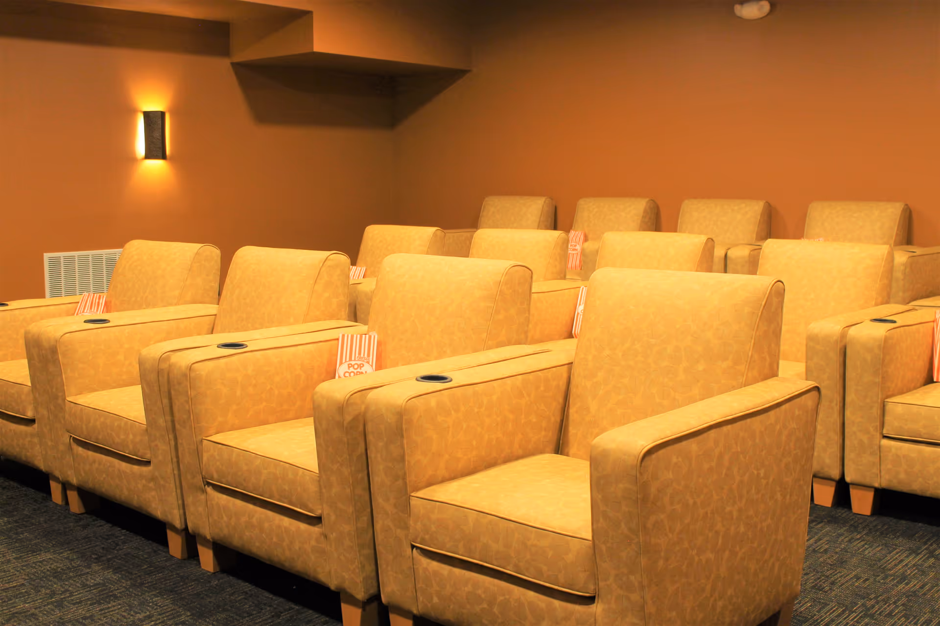 Small screening room with rows of beige upholstered armchairs with cup holders and popcorn bags.