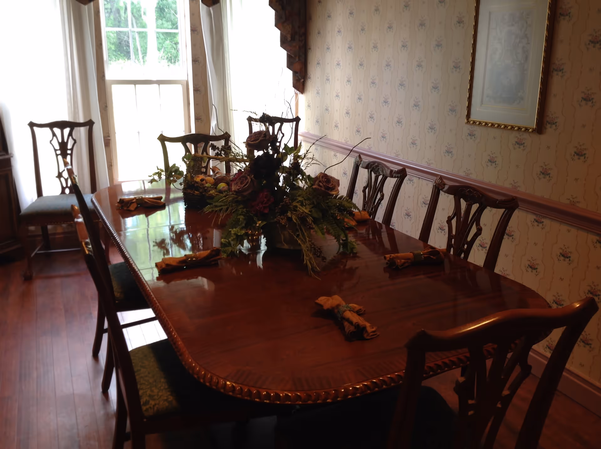 A traditional dining room with a polished wooden dining table surrounded by eight wooden chairs with green upholstered seats. The table is decorated with a floral centerpiece and has neatly folded napkins placed at each seat. The room has floral wallpaper, a framed picture on the wall, and a large window with white curtains letting in natural light.