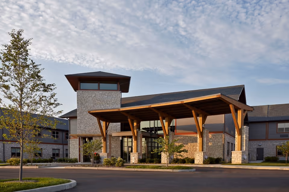 Exterior view of Wellbrooke of Westfield facility showing a modern building with stone and wood accents, a large covered entrance supported by wooden beams, and a clear sky with scattered clouds.