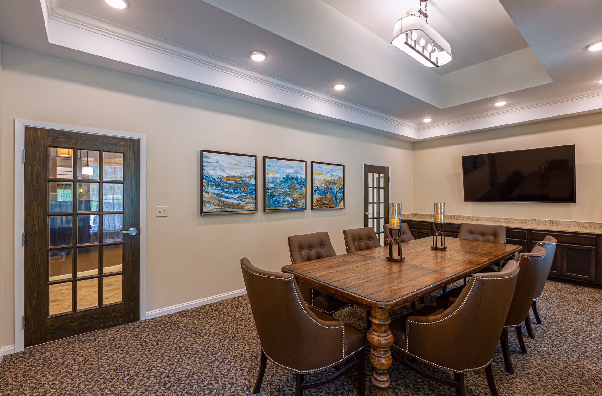 A conference or meeting room with a large wooden table surrounded by eight brown upholstered chairs. The room has beige walls, a patterned carpet, and a tray ceiling with recessed lighting and a central hanging light fixture. On one wall, there are three abstract paintings in blue and gold tones. A flat-screen TV is mounted on another wall above a long countertop with dark cabinetry. Two wooden doors with glass panels are visible.