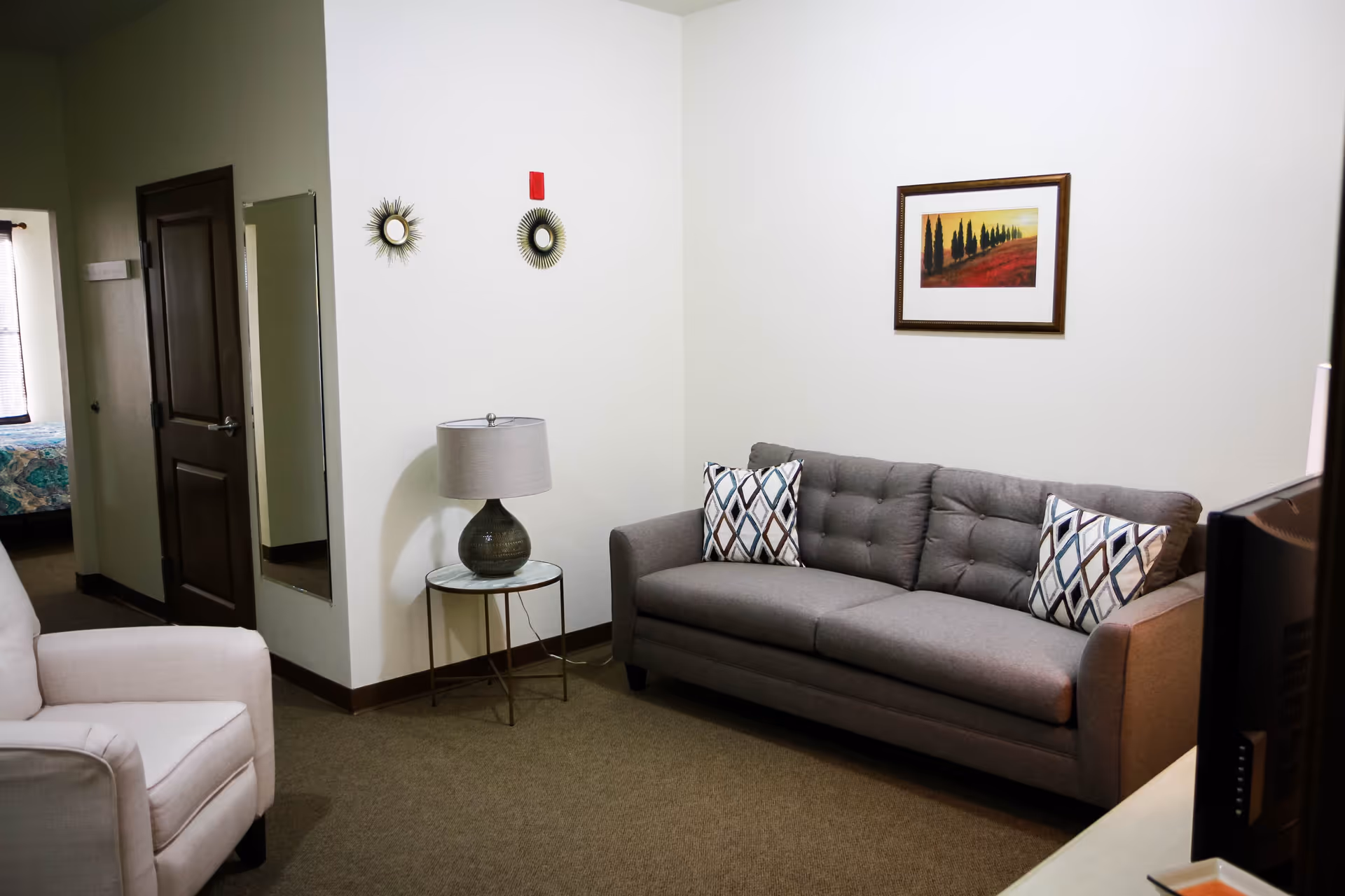 A cozy living room area with a gray sofa adorned with two patterned pillows, a white armchair, a small round side table with a lamp, two decorative round mirrors on the wall, and a framed landscape painting. A doorway leads to a bedroom with a bed covered in a blue and green patterned bedspread.