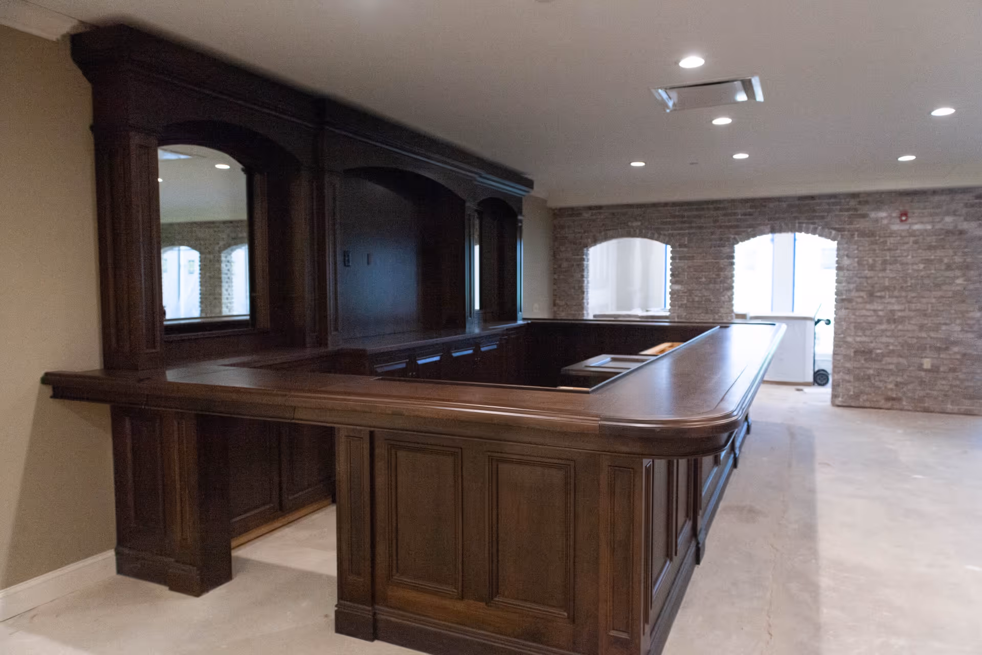 Large empty room featuring a dark wood U-shaped bar/counter and an exposed brick wall with arched windows.
