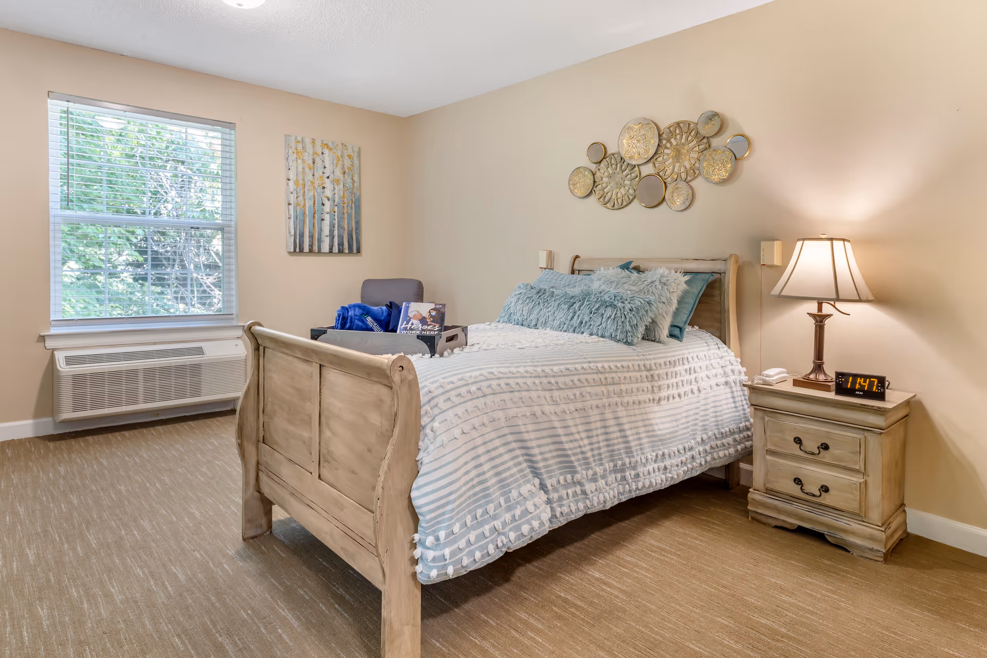 A cozy bedroom with a wooden bed frame, a bed made with a white and light blue striped comforter and fluffy blue pillows. Next to the bed is a wooden nightstand with a lamp and a digital clock showing 1:47. A window with blinds lets in natural light, and a chair with a blue blanket and a small tray is placed near the window. The walls are beige with decorative wall art above the bed.