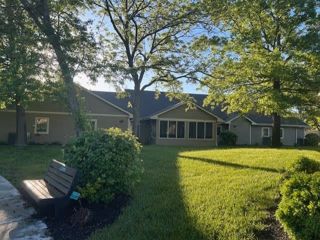A sunny outdoor scene at Aldersgate Village Life Plan Community showing a green lawn with trees casting shadows, a wooden bench on a paved path, and a single-story building with multiple windows in the background.