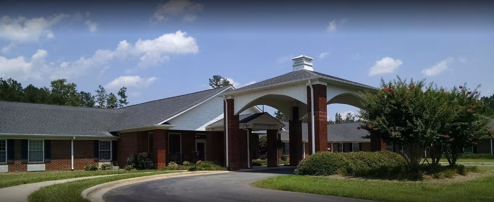 Single-story brick senior living building with a covered porte-cochere entrance, paved driveway and landscaped shrubs under a blue sky.