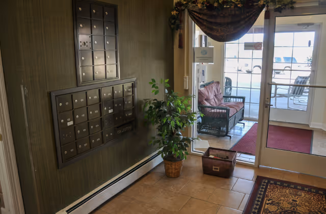 Interior view of a senior living facility entrance area with a wall-mounted mailbox unit, a potted plant, a basket on the floor, and glass doors leading outside to a patio with seating.