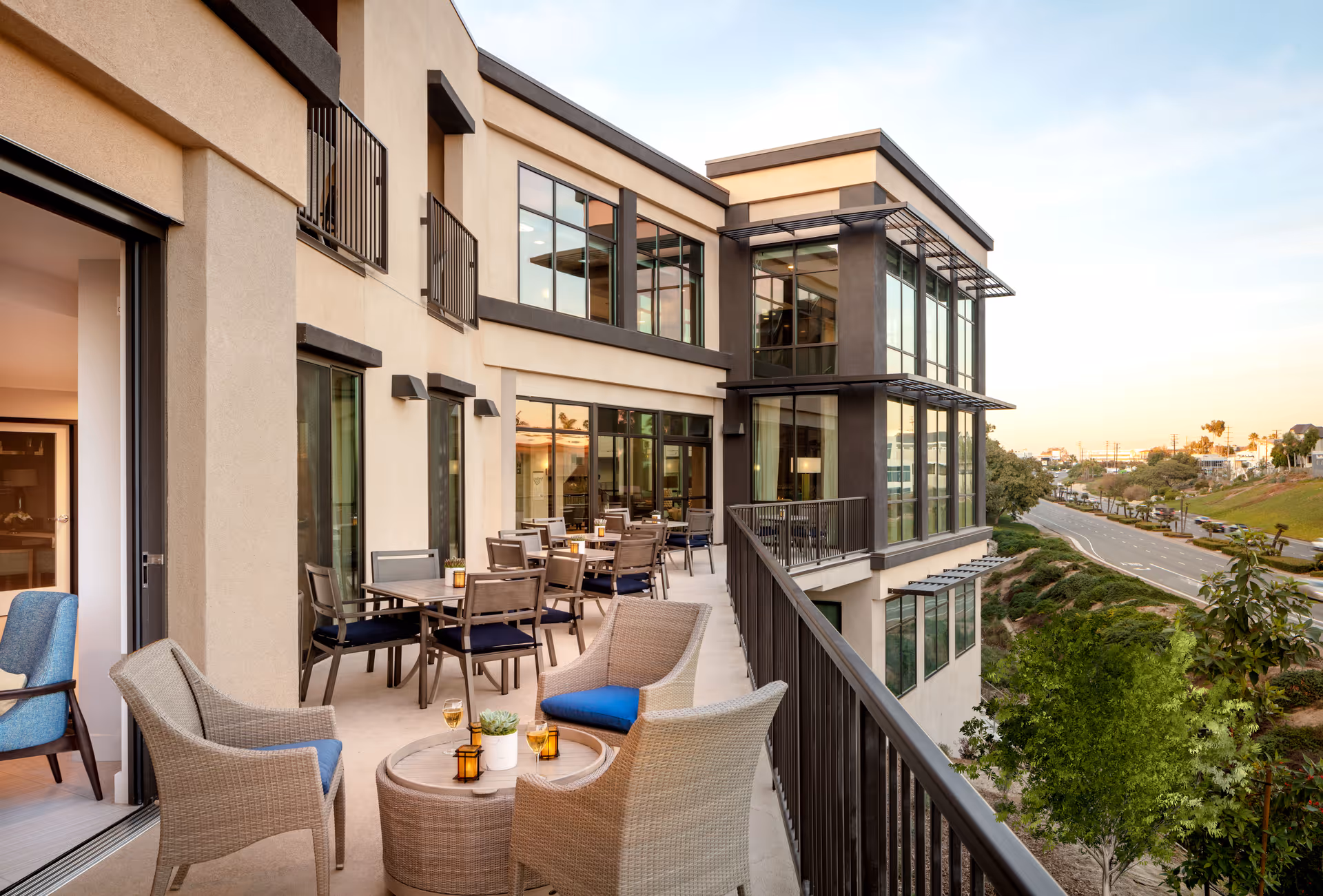 Outdoor balcony area at Atria Newport Beach with multiple seating arrangements including wicker chairs with blue cushions and tables. The balcony overlooks a road and greenery with a modern building facade featuring large windows.