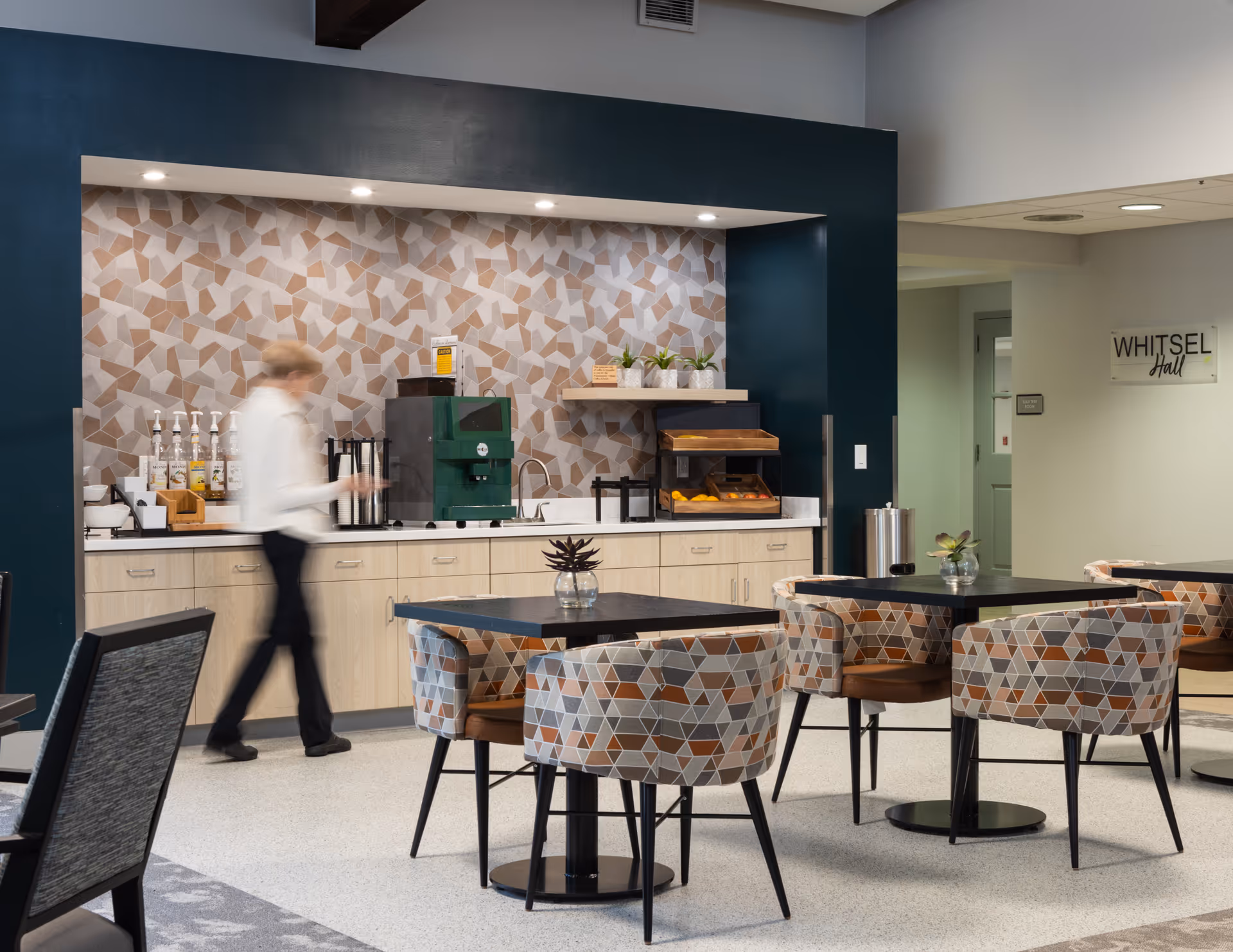 A modern dining area with patterned chairs around black tables. In the background, there is a counter with a coffee machine, syrup bottles, and a display case with fruit. A person is walking past the counter. The wall behind the counter has a geometric patterned backsplash and dark blue framing. A sign on the adjacent wall reads 'WHITSEL Hall'.
