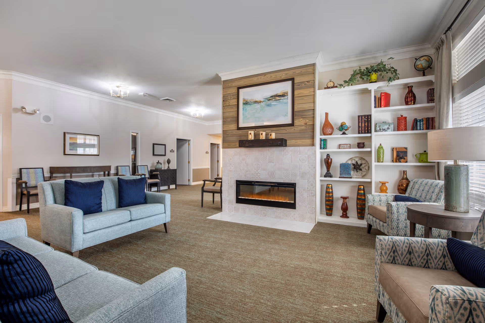 A bright and spacious living room area in a senior living facility featuring two light blue sofas with dark blue pillows, two patterned armchairs, a modern electric fireplace with a wooden accent wall above it, and built-in white shelves decorated with various vases, books, and plants. The room has beige carpeting, white walls, and large windows with blinds allowing natural light to enter.