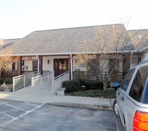 Exterior view of a single-story building with a sloped roof, featuring a wheelchair-accessible ramp leading to a covered entrance with double doors. There are leafless trees and shrubs near the entrance, and a silver SUV is parked in the foreground.