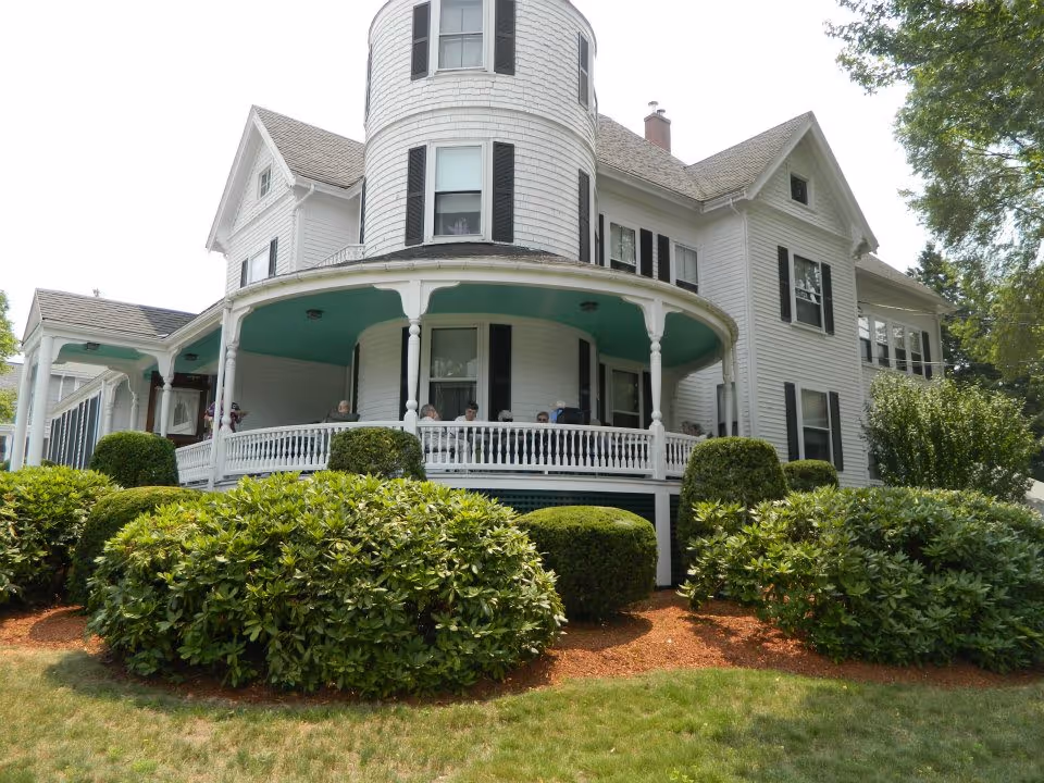 White Victorian-style house with a rounded turret and a covered wraparound porch, surrounded by manicured shrubs and lawn.