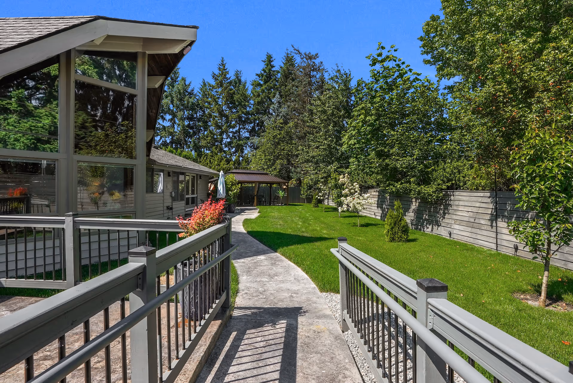 Outdoor view of a senior living facility with a concrete pathway leading through a green lawn bordered by trees and shrubs. A building with large windows and a gray railing is on the left side, and a covered seating area is visible in the background under a clear blue sky.