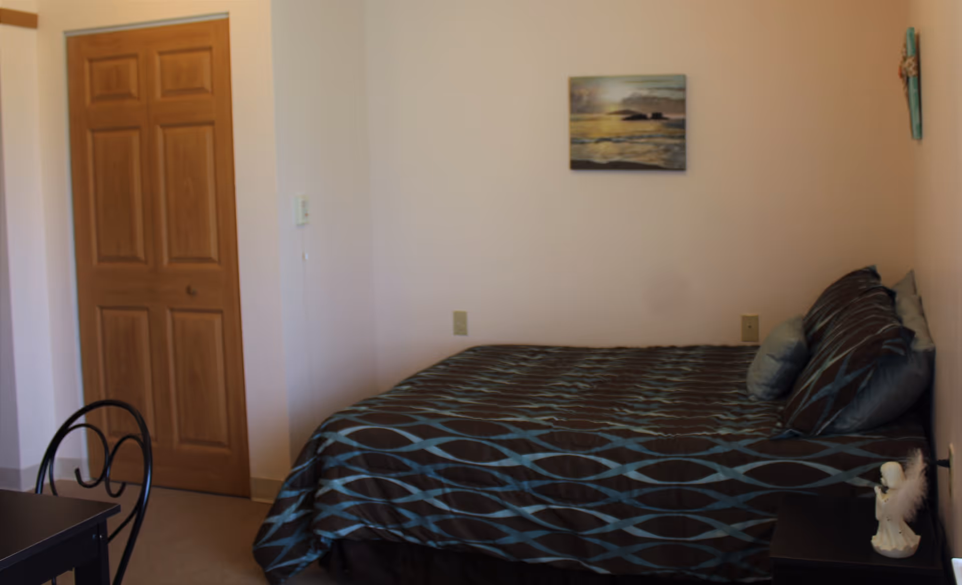 A bedroom with a bed covered in a dark blue and brown patterned bedspread, several pillows, a wooden door, a small black table with a black metal chair, a nightstand with a small angel figurine, and a painting of a sunset over the ocean on the wall.