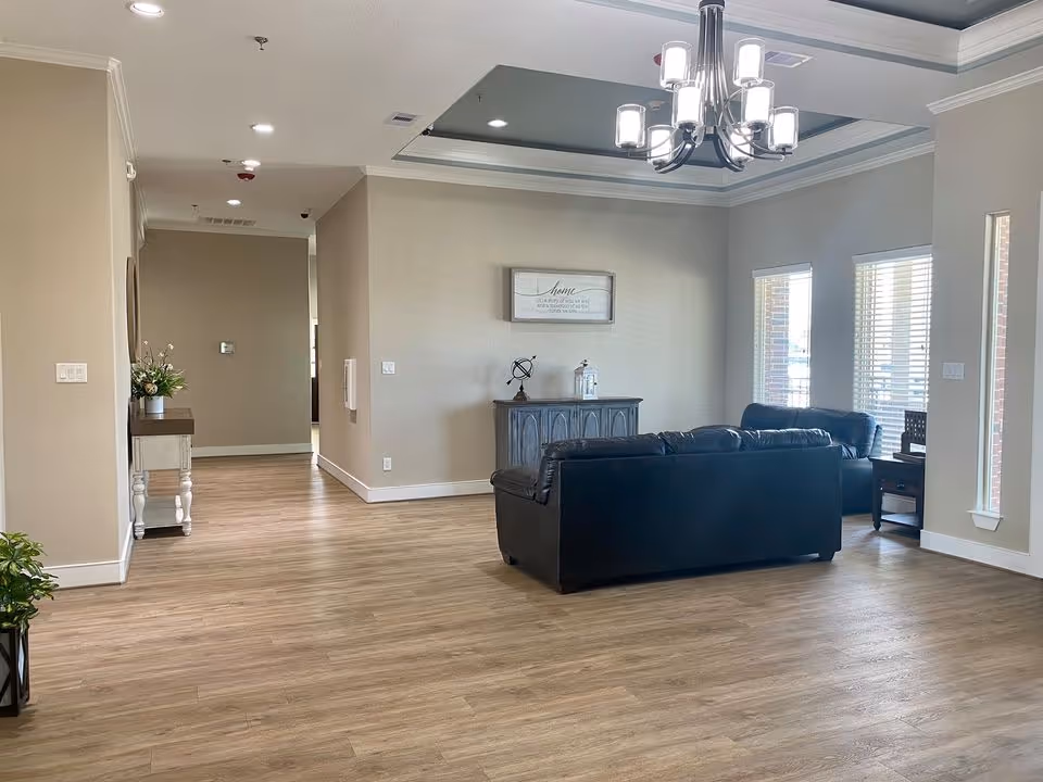 A spacious living room area with light wood flooring, beige walls, and a tray ceiling with a modern chandelier. The room features a black leather sofa and armchair arranged around a small wooden cabinet with decorative items. Large windows with blinds allow natural light to enter the room. A small table with flowers is visible in the hallway to the left.
