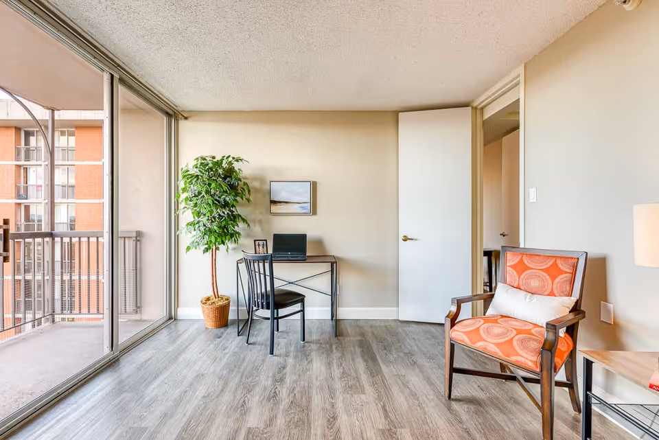 A bright room with a large sliding glass door leading to a balcony. Inside, there is a small desk with a laptop and a chair, a potted plant, an orange patterned armchair with a white cushion, and a side table with a lamp. The floor is wood laminate, and the walls are light-colored.