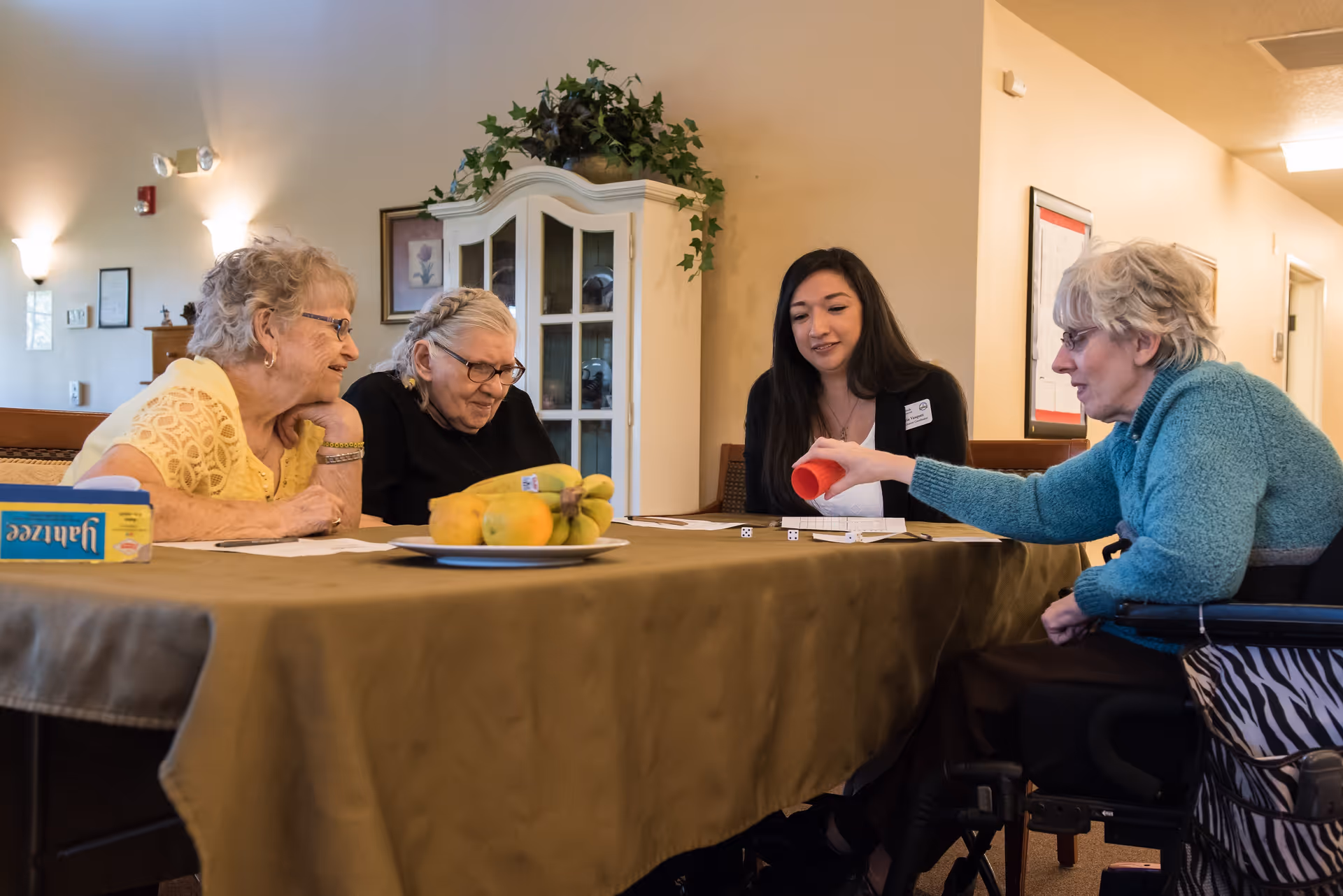 Three elderly women and a younger woman sitting around a table playing a dice game in a cozy room. One elderly woman in a wheelchair is rolling dice using a red cup. The table has a plate of bananas and other fruit, and a game box labeled 'Yahtzee'. The room has warm lighting, a cabinet with plants on top, and framed pictures on the wall.
