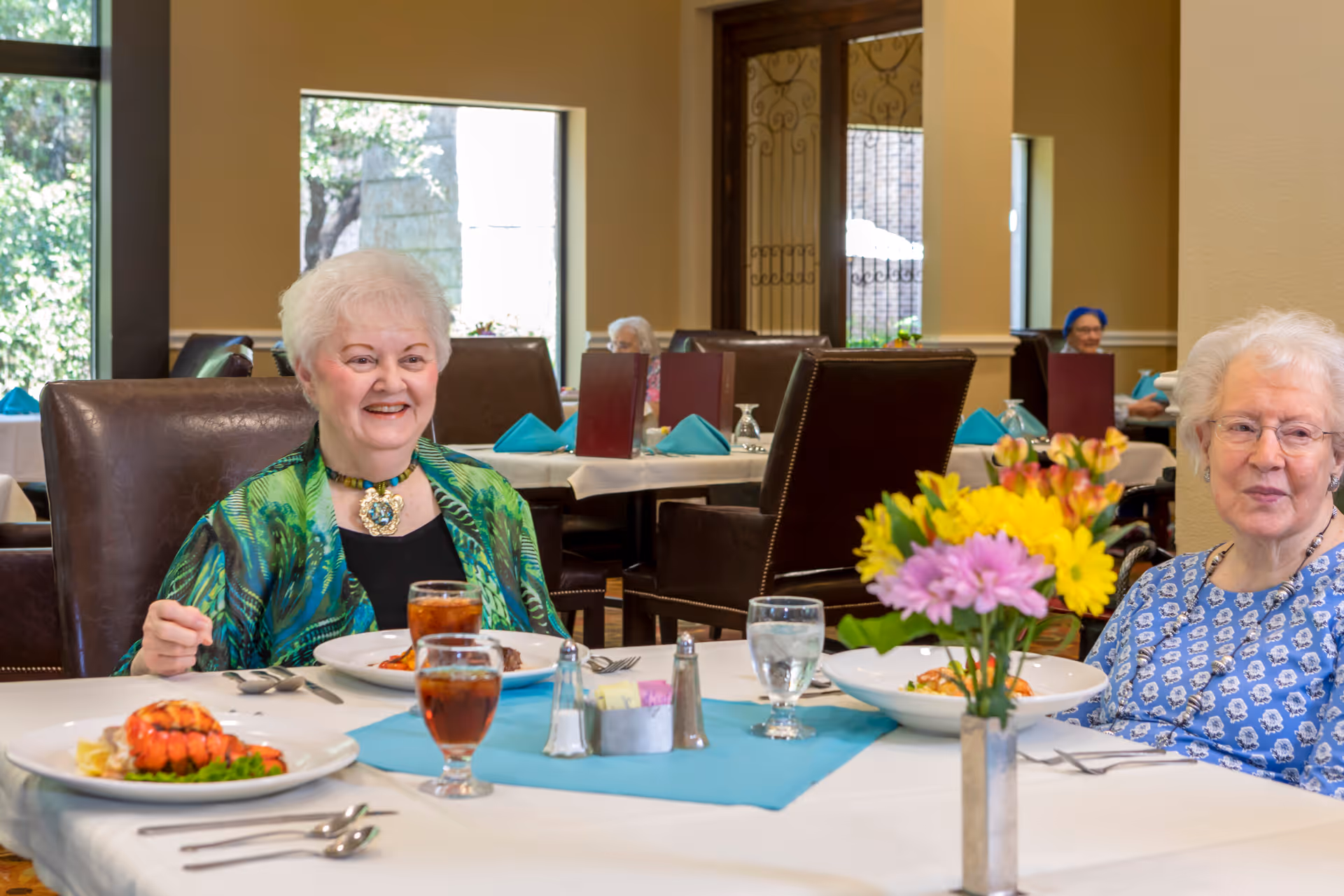 Two elderly women sitting at a dining table in a well-lit dining room, enjoying a meal with plates of food and glasses of iced tea in front of them. The table is decorated with a small bouquet of colorful flowers and has a blue table runner. Other tables with similar settings and menus are visible in the background.