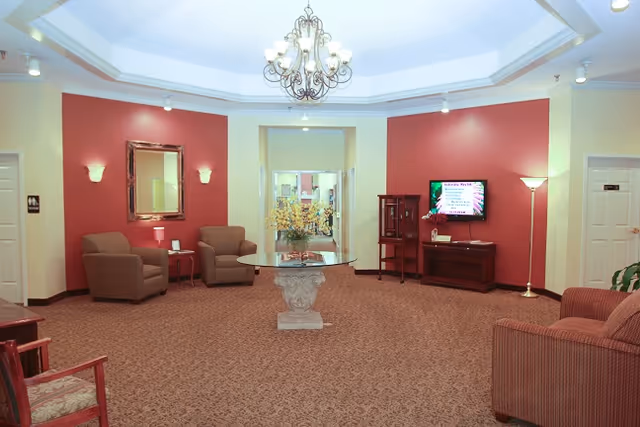 A spacious living room area with red accent walls and beige carpeting. The room features a round glass table with a floral centerpiece in the middle, two armchairs with a small side table and lamp between them, a wall-mounted TV displaying information, a wooden cabinet, a floor lamp, and a chandelier hanging from a tray ceiling. There are doorways and mirrors visible in the background.