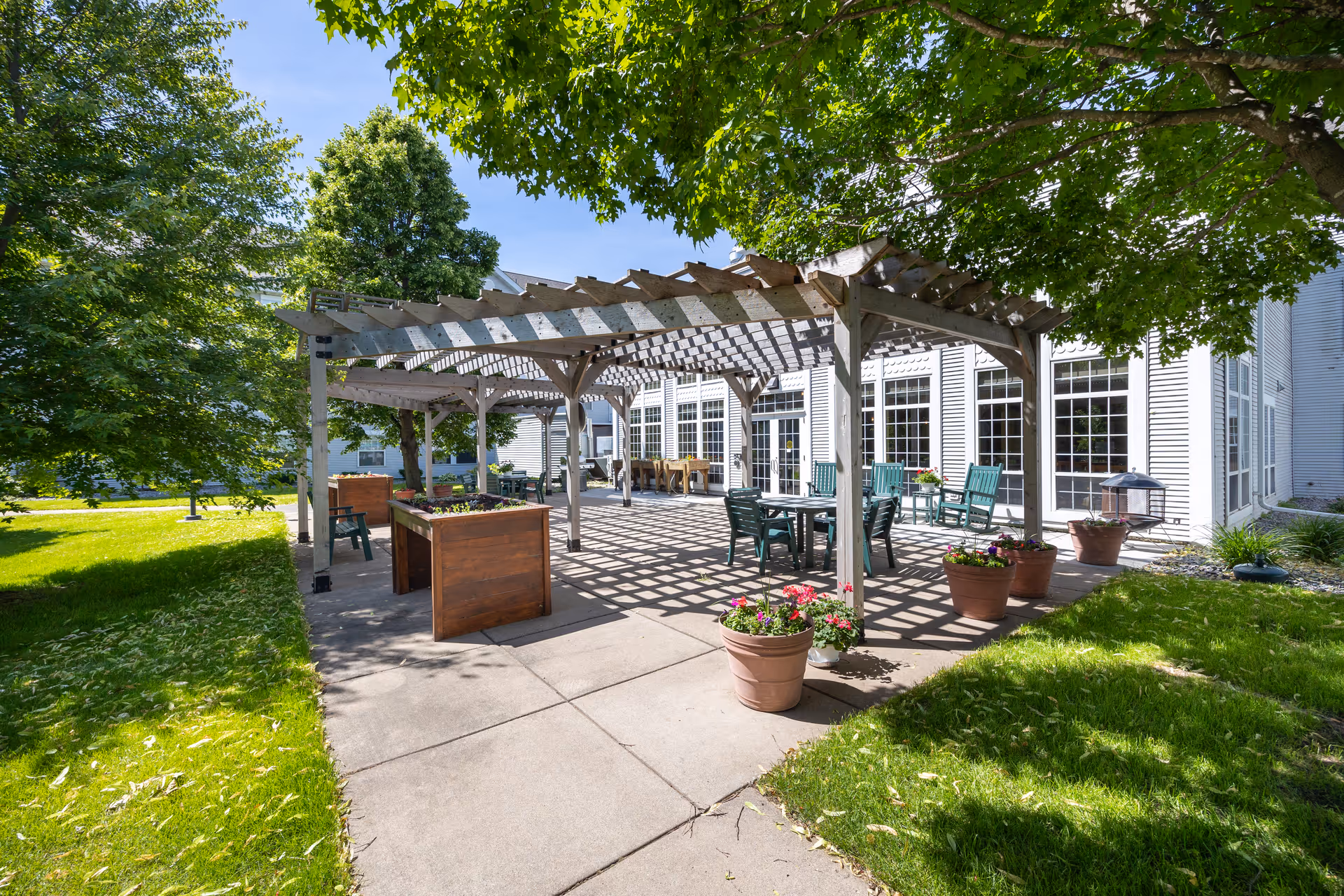Sunlit outdoor patio with a wooden pergola, potted plants, tables and chairs beside a white multi-windowed building.