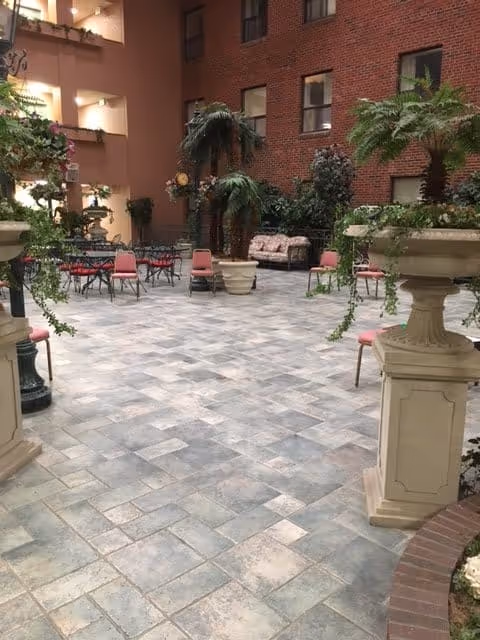 Indoor courtyard area with tiled flooring, several potted plants including large ferns, multiple pink chairs arranged around black metal tables, and a cushioned bench against a brick wall. The space is surrounded by building walls with windows and balconies.