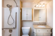 A clean and well-lit bathroom featuring a shower with a handheld showerhead and grab bar, a white toilet, a vanity with a granite countertop and sink, a wall-mounted cabinet with shutter-style doors, and a mirror with a light fixture above it.