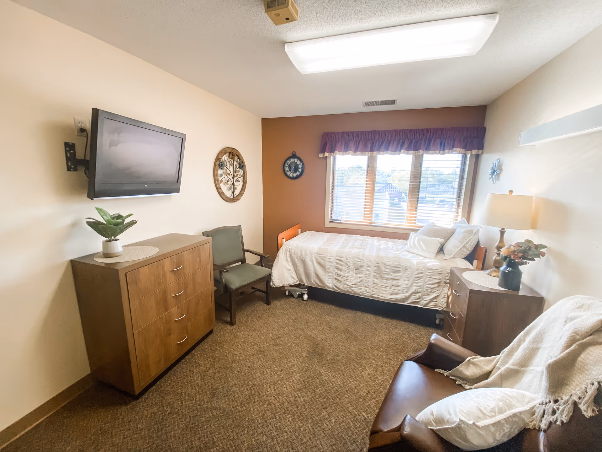 A cozy bedroom in a senior living facility with a single bed covered in white bedding positioned near a window with blinds and a purple valance. The room includes a wooden dresser with a potted plant on top, a green armchair, a brown leather armchair with a white throw blanket and pillow, a small wooden nightstand with a lamp and a vase of flowers, and a wall-mounted flat-screen TV. The walls are painted in light beige and one accent wall is a warm brown color.