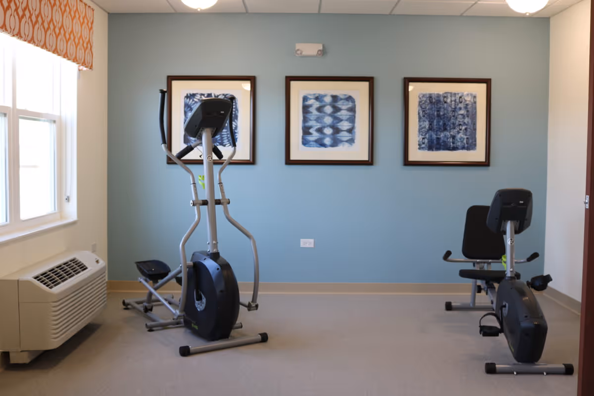 A small exercise room with two pieces of cardio equipment, an elliptical machine and a recumbent bike, positioned on a light gray floor. The walls are painted light blue and decorated with three framed abstract blue artworks. A window with an orange patterned valance is on the left side, and a white air conditioning unit is below the window.