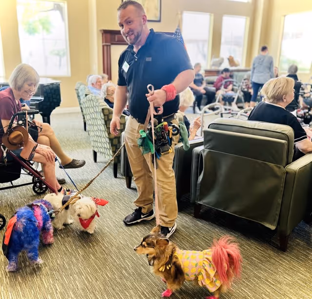 A man holding leashes of three small dogs dressed in colorful outfits interacts with elderly residents seated in a common area with armchairs and large windows letting in natural light.