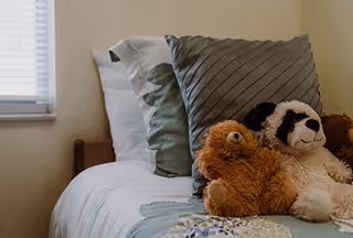 A neatly made bed with multiple pillows including a gray striped pillow and a green pillow, along with two stuffed animals, a brown teddy bear and a black and white panda, placed on the bed near a window with blinds.