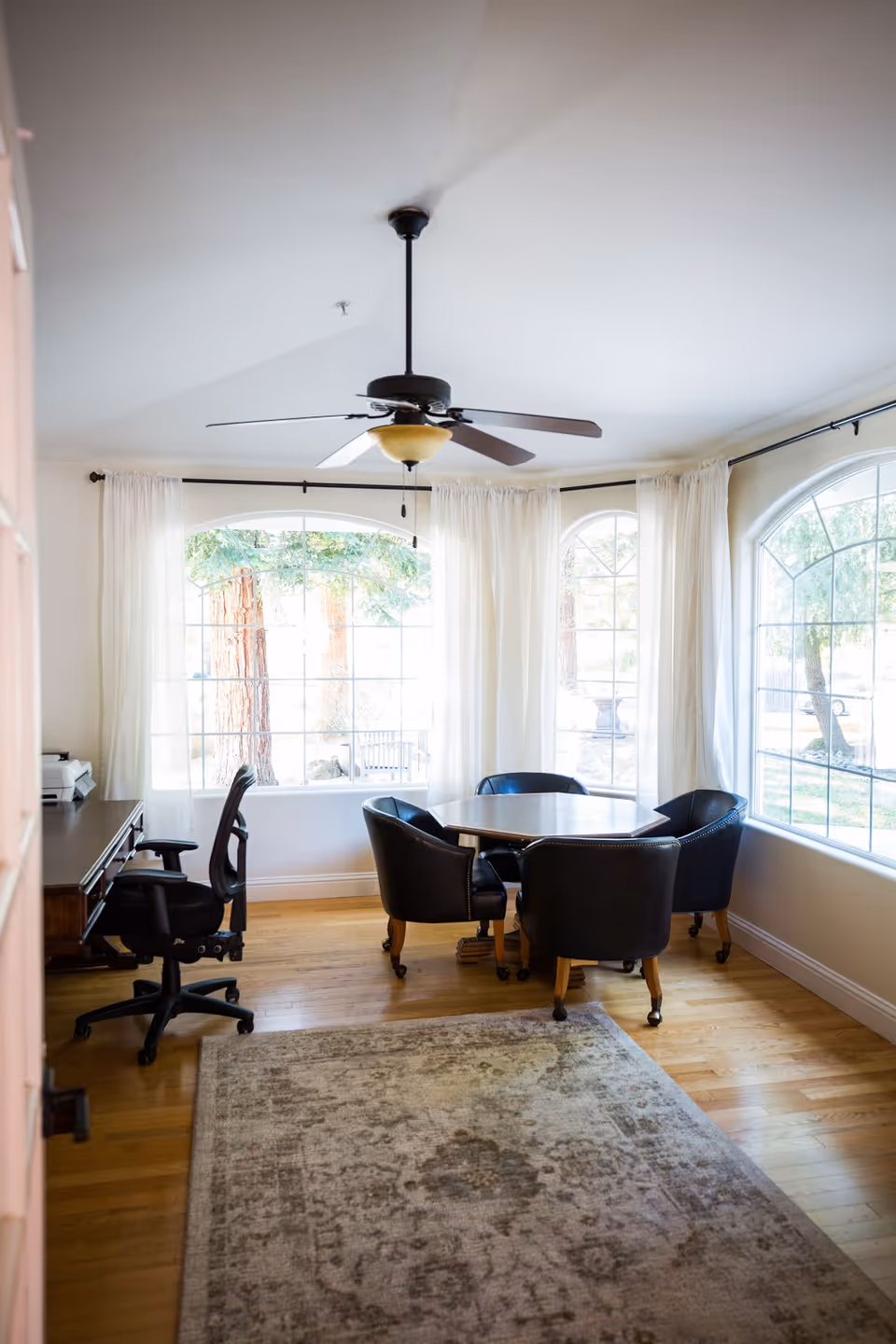A bright room with large arched windows covered with sheer white curtains, a ceiling fan with light, a round wooden table surrounded by four black leather chairs on casters, a wooden desk with an office chair, and a patterned area rug on a hardwood floor.