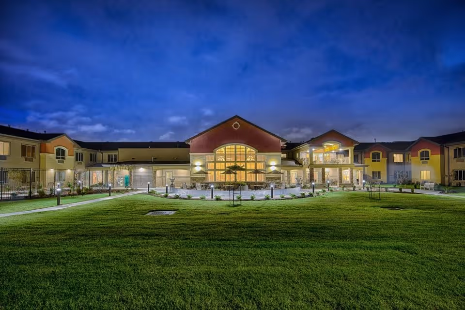 Nighttime exterior view of a large assisted living facility building with well-lit windows and outdoor lighting. The building is surrounded by a neatly maintained green lawn and paved walkways.