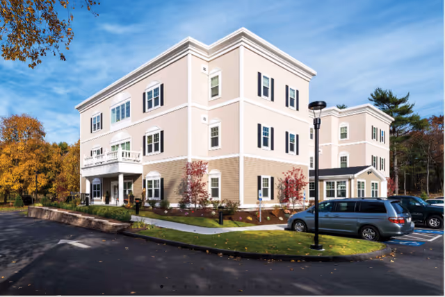 Exterior view of Windrose at Weymouth, a senior living facility, with cars and trees.