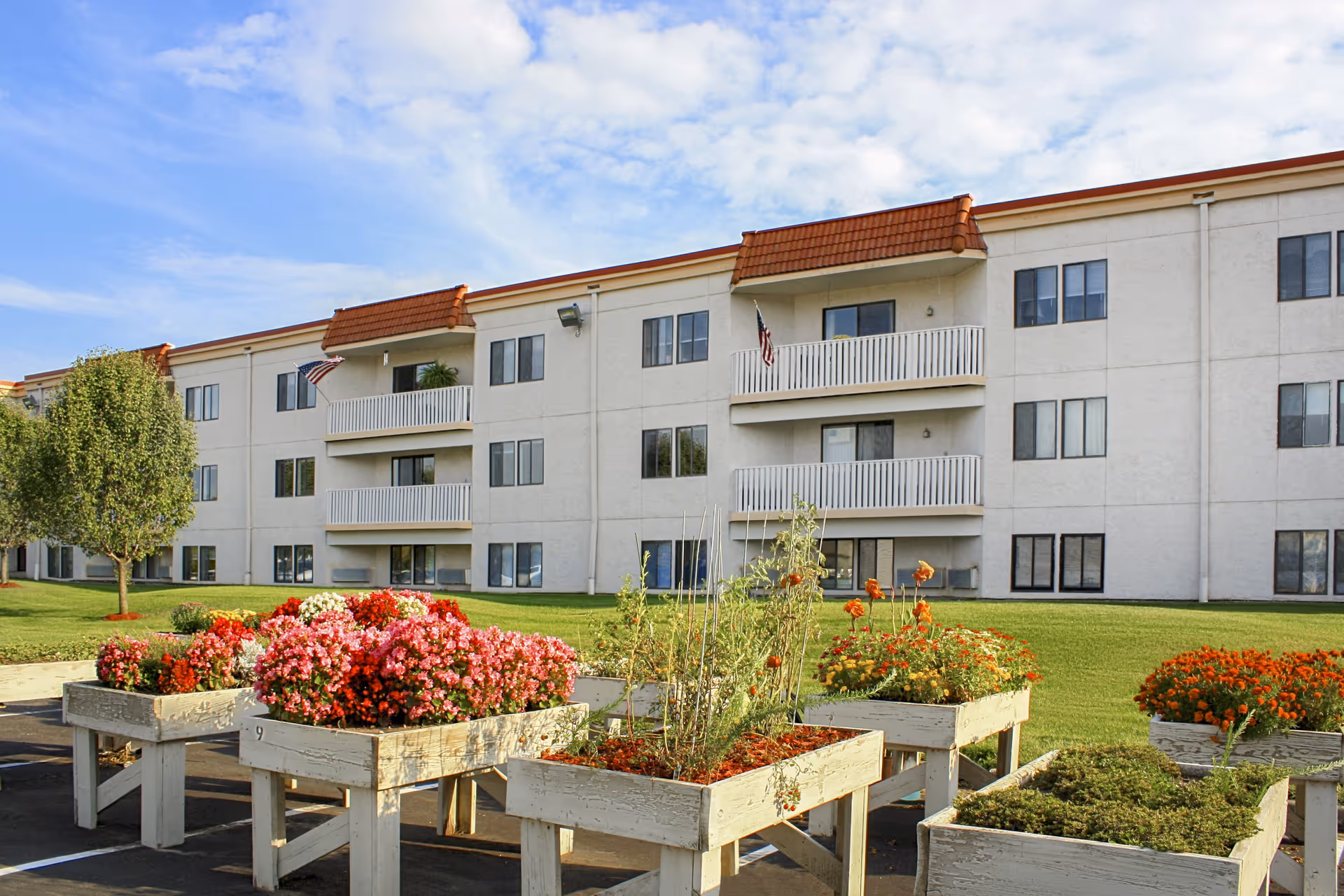 Exterior view of a three-story senior living facility building with balconies and American flags. In the foreground, there are raised garden beds filled with colorful flowers and plants, set on a paved area with a green lawn and a tree nearby under a partly cloudy sky.