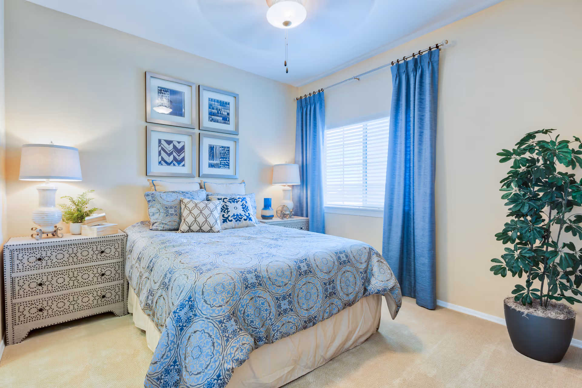Bright bedroom with a made bed featuring patterned blue bedding, two bedside tables with lamps, framed wall art, blue curtains, and a potted plant.