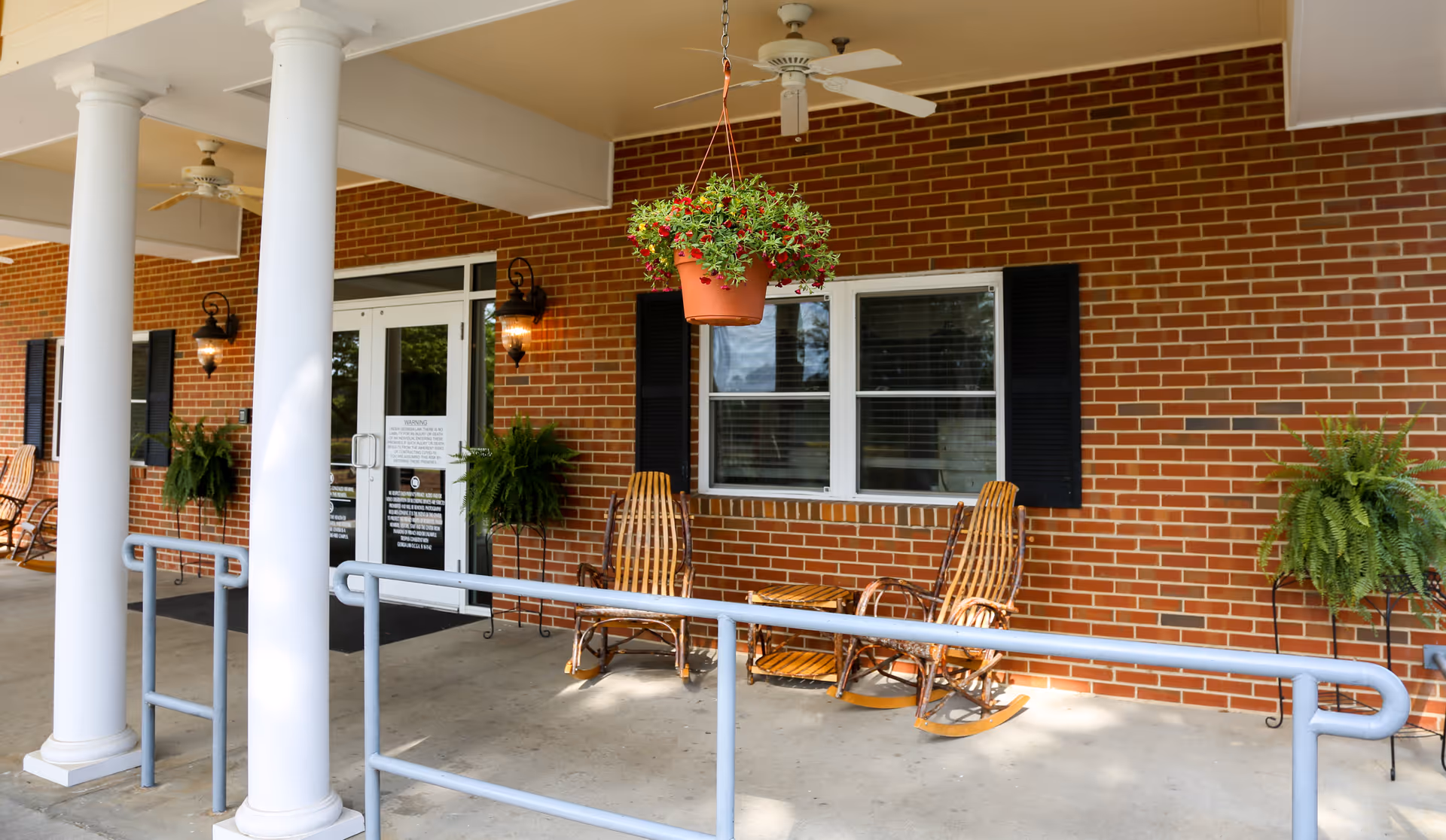 Covered outdoor patio area with white columns, hanging flower pot, two wooden rocking chairs, potted ferns, and double glass doors leading inside a brick building.