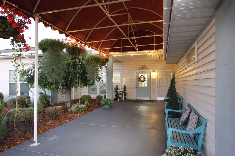 Covered outdoor entrance area of a senior living facility with a red canopy overhead, teal benches with decorative pillows along the right wall, potted plants, hanging flower baskets, and a door decorated with a wreath at the far end.