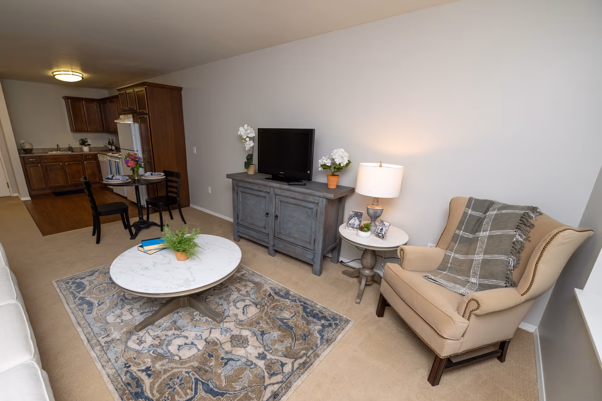 A cozy living area with a beige armchair draped with a plaid throw blanket, a round side table with a lamp and small plants, a blue-gray TV stand with a flat-screen TV and potted flowers, a round marble coffee table with a small plant and books, and a patterned area rug. In the background, there is a small dining table with two chairs and a kitchen with wooden cabinets and appliances.