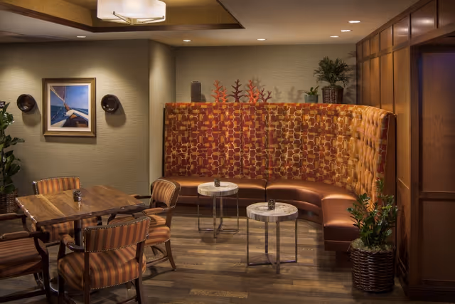 Warm, well-lit seating area featuring a curved upholstered banquette, small round side tables, a wooden dining table and striped chairs.