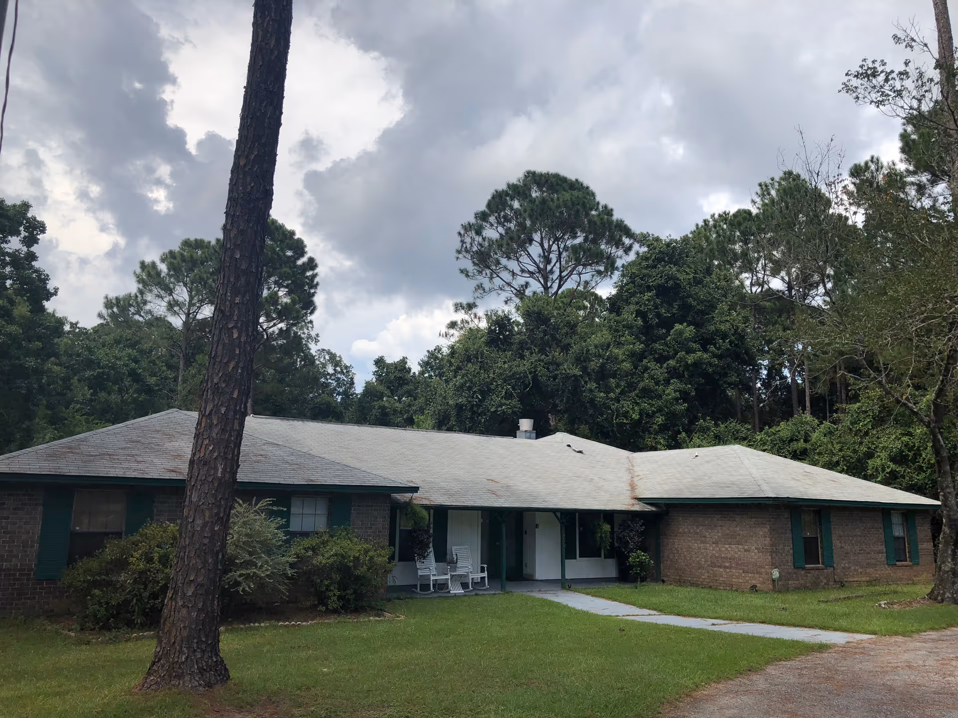Single-story brick building with a gray roof surrounded by trees and greenery under a cloudy sky. There are two white rocking chairs on the front porch and a paved walkway leading to the entrance.