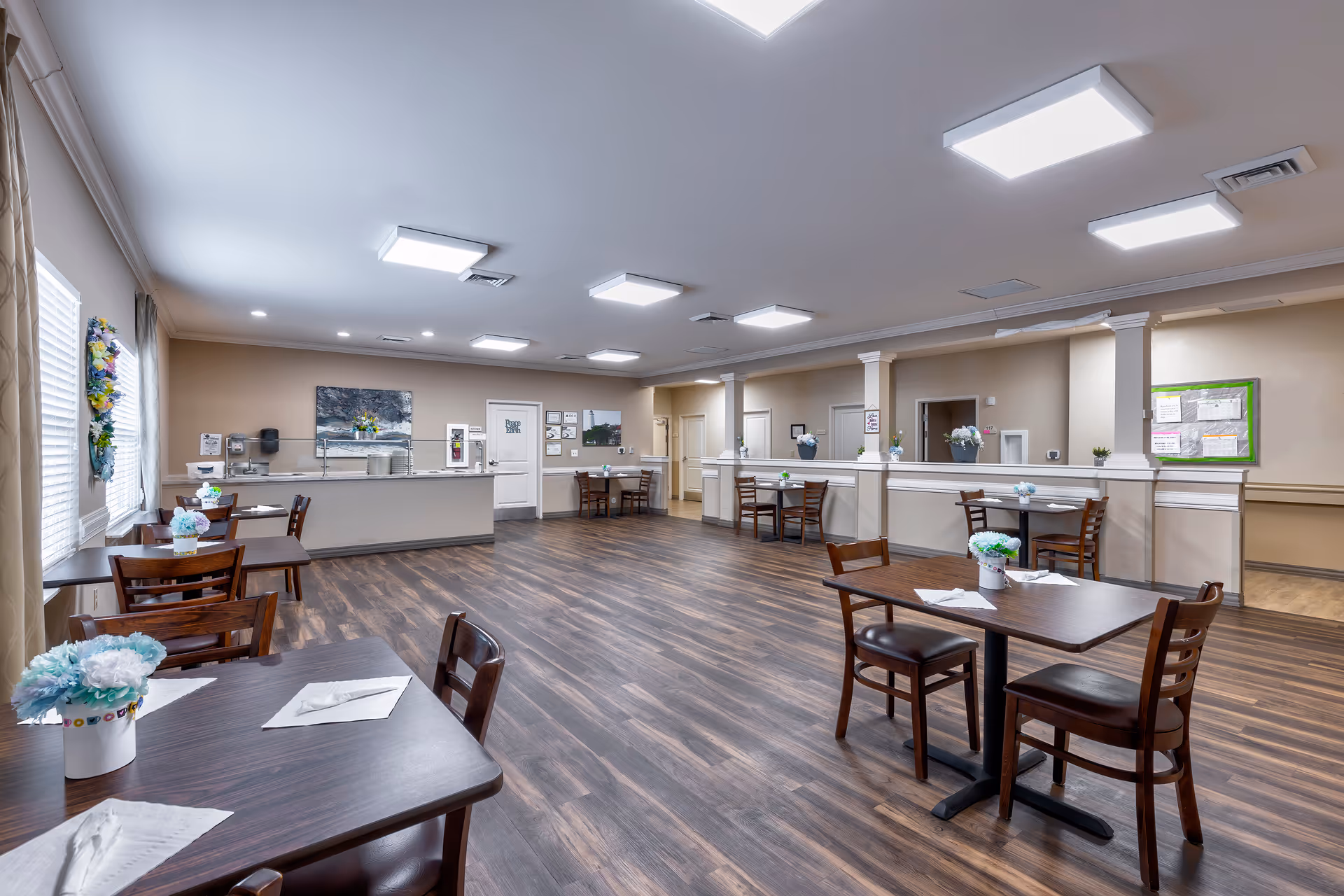 A spacious dining room with multiple wooden tables and chairs arranged neatly. Each table has a small flower arrangement and napkins. The room has wood flooring, beige walls, and bright ceiling lights. There is a counter area at the back with a coffee machine and other amenities. Windows with blinds are on the left side, and some wall decorations and bulletin boards are visible.