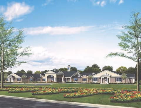 Exterior view of Benton House of Olathe, a single-story senior living facility with a large front lawn featuring landscaped flower beds and trees under a partly cloudy blue sky.